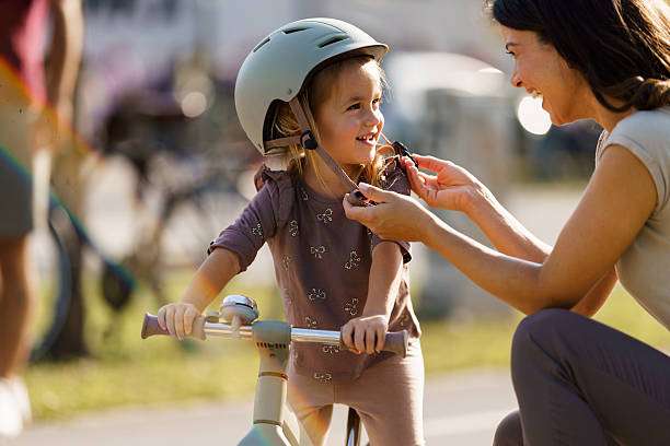Mom is helping a smiling girl put on a bike helmet before riding her balance bicycle.