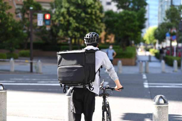 Delivery person wears black bike helmet and backpack, pushing bicycle on city street.